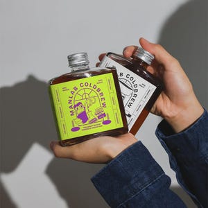 Peut inclure: Deux bouteilles carrées en verre de café infusé à froid avec des bouchons argentés sont tenues. L'une a une étiquette vert vif avec "MANLAB COLDBREW" et un dessin animé. L'autre a une étiquette blanche avec "B COLDBREW".