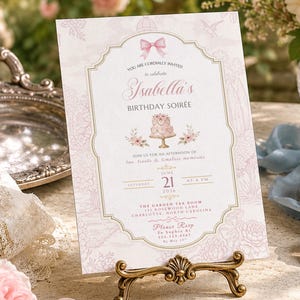May include: A pink and gold invitation for "Isabella's Birthday Soirée" displayed on a decorative gold stand. The invitation features a bow, cake, and floral elements. The text includes the date, time, and location.