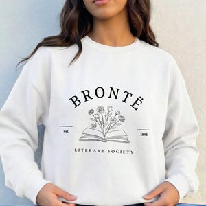 Puede incluir: Una sudadera blanca de manga larga y cuello redondo con un estampado gráfico en negro. El diseño incluye el texto "BRONTË LITERARY SOCIETY" con "est. 1893" y una ilustración central de un libro abierto con un ramo de flores saliendo.
