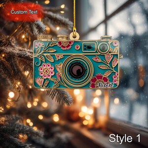 Peut inclure: Ornement de Noël en forme d'appareil photo, de couleur turquoise et dorée, avec des détails floraux et le nom "Robert". L'ornement est orné d'une ficelle dorée pour l'accrocher. Le motif de l'appareil photo comprend des fleurs roses et des feuilles. Le texte "Style 1" est en bas.
