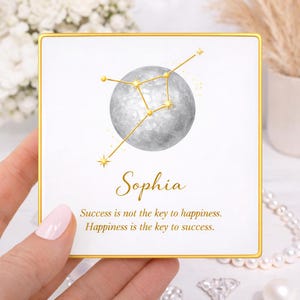 May include: A square, white card with a gold border features a watercolor illustration of a gray moon and gold constellation. The name "Sophia" is written in elegant script, with the quote "Success is not the key to happiness. Happiness is the key to success."