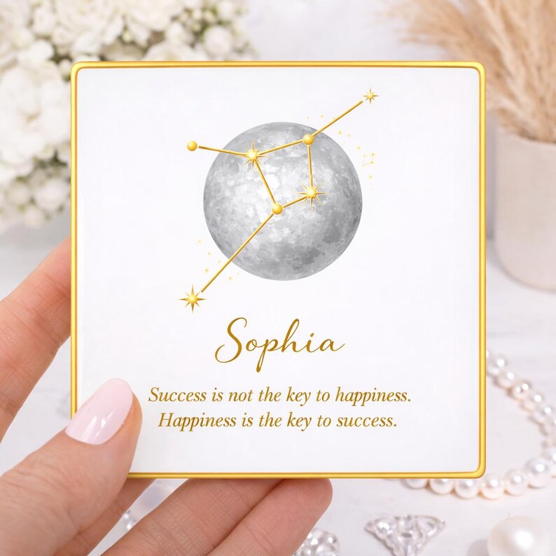 May include: A square, white card with a gold border features a watercolor illustration of a gray moon and gold constellation. The name "Sophia" is written in elegant script, with the quote "Success is not the key to happiness. Happiness is the key to success."