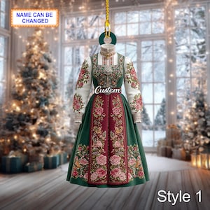Puede incluir: Adorno con un diseño de vestido tradicional. El vestido tiene un corpiño blanco con bordados florales, mangas largas y una falda verde. El diseño incluye la palabra "Custom" y "Style 1". El adorno cuelga de una cuerda dorada.