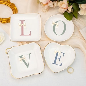 May include: Four white ceramic trinket dishes spelling "LOVE". Each dish has a gold rim, a letter in a different colour, and a bow. The "L" is square, the "O" is round, the "V" is wavy, and the "E" is heart-shaped.