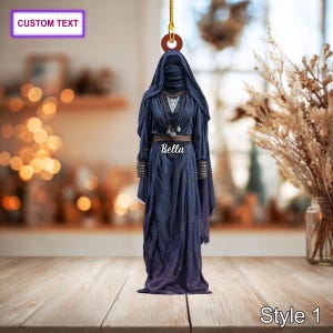 May include: A navy blue ornament depicting a hooded figure in a flowing gown. The figure has a veil covering the face and is adorned with necklaces and a belt. The name "Bella" is written on the garment. The ornament is hanging from a gold string. Text at the top says "CUSTOM TEXT" and at the bottom "Style 1".