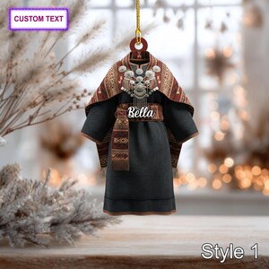 May include: Ornament featuring a stylized traditional outfit. The outfit is primarily black with a patterned shawl in shades of red, brown, and cream. The name "Bella" is displayed on the waist. The ornament has a gold hanging cord.