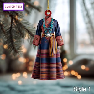 Peut inclure: Ornement représentant une robe traditionnelle bleue avec des détails rouges et dorés. La robe a une longue jupe rayée et des manches décoratives. Un collier turquoise et une ceinture avec le nom "Bella" sont visibles. L'ornement est étiqueté "Style 1" et contient le texte "CUSTOM TEXT".