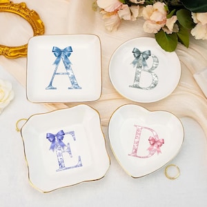 Könnte beinhalten: Vier weiße Keramik-Schmuckschalen mit goldenen Rändern. Jede Schale zeigt einen Buchstaben des Alphabets mit einer Schleife. Die Buchstaben sind A, B, D und E. Der Buchstabe A ist blau, B ist silber, D ist rosa und E ist lila.