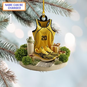 May include: A yellow sports jersey with the number 20, a pair of yellow running shoes, and a water bottle. The ornament includes a small road, gift boxes, and green bushes. The text "NAME CAN BE CHANGED" is visible.