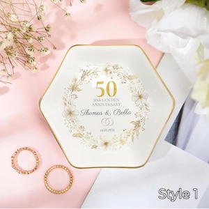 Puede incluir: Un plato de cerámica hexagonal blanco con borde dorado. El plato está decorado con texto dorado y diseños florales, conmemorando un 50 aniversario de bodas. El texto dice "50 50th Golden Anniversary Thomas & Bella 14.05.1976". Dos anillos dorados están junto al plato.