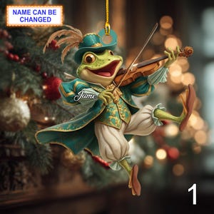 May include: A whimsical Christmas ornament featuring a frog playing a violin. The frog is dressed in a teal and gold outfit with a matching hat. The ornament has the text "Name Can Be Changed" and the name "Jame" on it.
