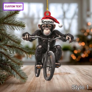 May include: A Christmas ornament featuring a chimpanzee riding a bicycle. The chimpanzee wears a Santa hat with the name "Bruno". The ornament has a gold string for hanging and the text "CUSTOM TEXT" is in the upper left corner. The words "Style 1" are in the lower right corner.