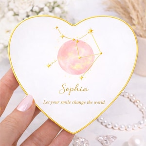 May include: A heart-shaped white trinket dish with a gold trim. The dish features a pink watercolor moon and constellation design, with the name "Sophia" and the phrase "Let your smile change the world."