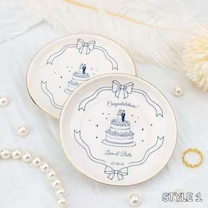 Puede incluir: Dos platos de cerámica blanca con borde dorado, con una ilustración azul de una tarta de boda y una pareja. Los platos tienen un diseño de cinta y el texto "¡Felicidades!" y nombres. Un anillo y perlas están en el fondo.