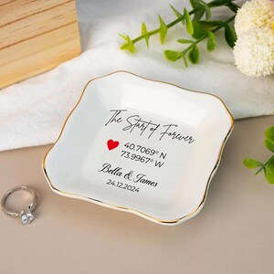 May include: A white ceramic ring dish with a gold rim. The dish has the text "The Start of Forever", a red heart, coordinates, and the names "Bella & James" with a date. A diamond ring sits to the left.