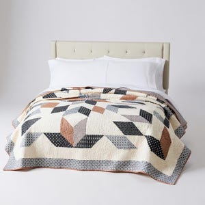 Könnte beinhalten: Eine cremefarbene Steppdecke mit geometrischen Mustern in Braun-, Schwarz- und Grautönen. Die Steppdecke hat einen Rand mit einem sich wiederholenden grau-weißen Design. Sie ist über einem Bett mit weißen Laken und Kissen drapiert. Maße in Zentimetern.