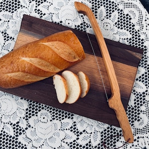 May include: A loaf of golden-brown bread, partially sliced, rests on a wooden cutting board. A wooden bread slicer with a wire blade is positioned nearby. The board has a two-tone wood grain design.