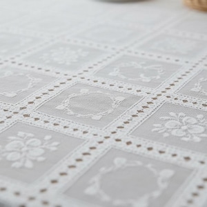 Könnte beinhalten: Weißes Spitzentischtuch mit einem sich wiederholenden quadratischen Muster. Jedes Quadrat weist ein Blumen- oder Kreisdesign auf. Die Ränder der Quadrate sind mit einem gepunkteten Muster umrandet. Das Tischtuch besteht aus einem zarten, transparenten Stoff.