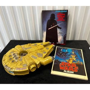 Puede incluir: Un juguete amarillo del Halcón Milenario se encuentra junto a dos libros de Star Wars. Un libro presenta a Darth Vader, y el otro es un álbum de Star Wars con una portada azul. El juguete es una réplica detallada de la nave espacial.