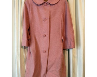May include: A vintage, pale pink coat featuring a rounded collar and three buttons. The coat has three-quarter length sleeves and a tailored fit. It is suspended on a wooden hanger. The background is a neutral colour.