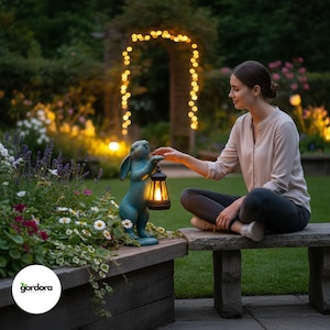 Könnte beinhalten: Eine türkisfarbene Hasenstatue, die eine leuchtende Laterne hält. Die Statue steht in der Nähe eines Blumenbeets mit weißen und lila Blumen. Eine Person sitzt auf einer Steinbank und berührt die Laterne. Im Hintergrund ist ein Garten mit Lichterketten zu sehen.