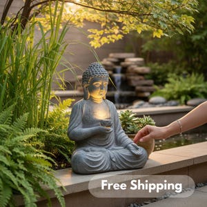 May include: A gray stone Buddha statue in a seated meditation pose, holding a small bowl. The statue is illuminated from within, set in a garden with greenery and a small waterfall. The text "Free Shipping" is at the bottom.