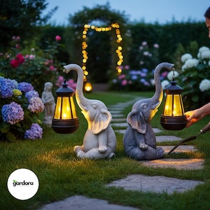 May include: Two elephant-shaped garden statues, one light gray and one dark gray, each holding a lantern with a warm yellow glow. The statues are set on a grassy lawn, with flowers and a string of lights in the background.
