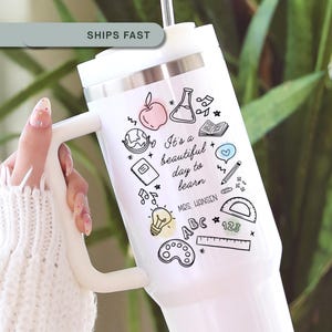 May include: White insulated tumbler with a handle, featuring a black and pastel-colored illustration of school-related icons and the text "It's a beautiful day to learn." The tumbler includes the name "Mrs. Hansen".