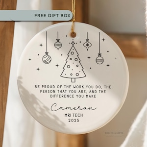 May include: A round, off-white ceramic Christmas ornament with a gold string. The ornament features a black line drawing of a Christmas tree and hanging ornaments. Text includes "Be proud of the work you do..." and "Cameron MRI TECH 2025."
