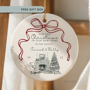 May include: A round, cream-colored Christmas ornament with a red ribbon design and the text "OUR FIRST Christmas IN OUR NEW HOME." It features a sketch of a fireplace, Christmas tree, and the names "Conrad & Belly" with the year 2025.