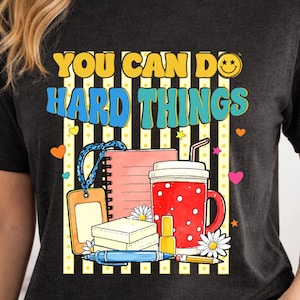 May include: A charcoal grey t-shirt featuring a graphic print with the text YOU CAN DO HARD THINGS. The design includes a spiral notebook, a red polka-dot mug, a yellow ID badge, a blue pen, a yellow pencil, and white daisy flowers.