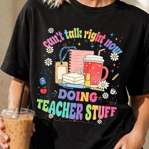 May include: A black short-sleeve t-shirt featuring a colourful graphic with the text "can't talk right now! DOING TEACHER STUFF" surrounded by illustrations of a notebook, a red mug, a pen, a pencil, a small apple, and white daisy flowers.