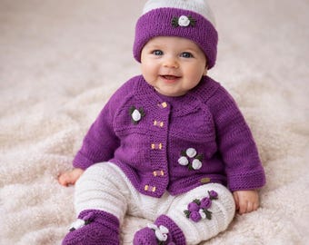 Op de afbeelding: Een gebreide baby-set in paars en wit, bestaande uit een vest met lange mouwen, een bijpassende broek, een muts met een pompon en slofjes. De set is voorzien van kleine witte bloemapplicaties met groene blaadjes op de muts, het vest en de broek.