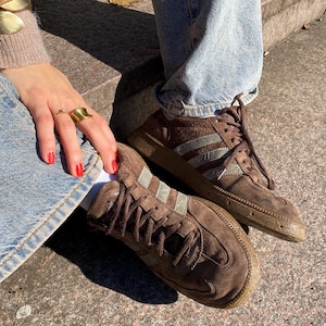 Könnte beinhalten: Braune Wildleder-Sneaker mit silbernen Streifen und Schnürsenkeln. Die Schuhe sind abgenutzt und liegen auf einer Steinoberfläche. Das Bild zeigt auch eine Person, die blaue Jeans und eine braune Lederjacke trägt.