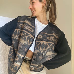 Peut inclure: Veste boutonnée avec un motif géométrique dans les tons de gris, noir et beige. La veste a des manches noires et une poche zippée. Le modèle porte un haut blanc et un pantalon beige.