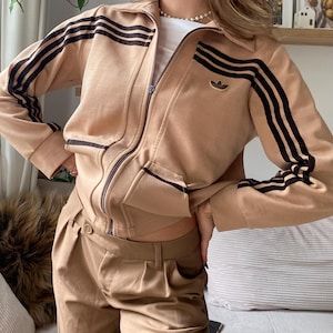 Könnte beinhalten: Beigefarbene Trainingsjacke mit Reißverschluss und schwarzen Streifen auf den Ärmeln sowie einem kleinen Logo auf der Brust. Dazu eine passende beige Hose. Vintage-Look.