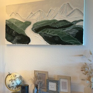 May include: An abstract landscape painting featuring a river winding through green hills and white mountains under a light gray sky. The artwork is mounted on a white wall above a white table with decorative items.