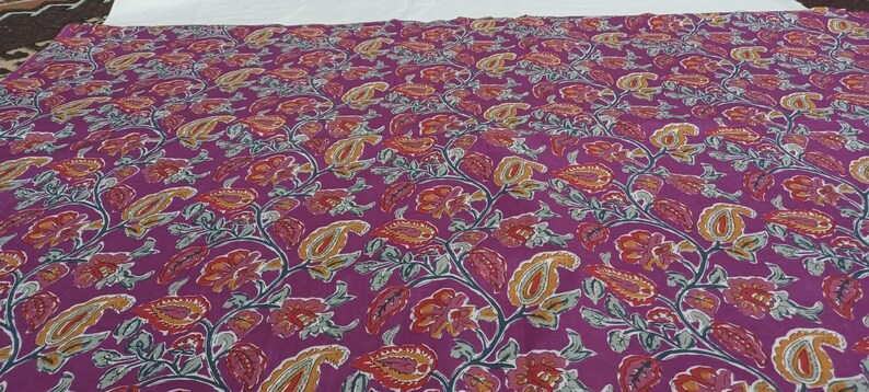 May include: A large piece of fabric with a repeating floral pattern. The design features paisley and floral elements in shades of red, orange, yellow, and gray against a deep purple background. The fabric appears to be cotton or a similar material.