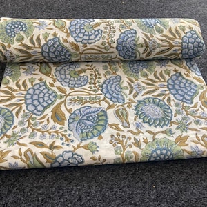 May include: A folded and rolled fabric with a floral pattern. The design features large blue and green flowers with brown stems and leaves on a cream background. The fabric is displayed on a dark gray surface.