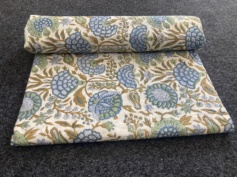 May include: A folded and rolled fabric with a floral pattern. The design features large blue and green flowers with brown stems and leaves on a cream background. The fabric is displayed on a dark gray surface.