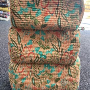 May include: Three stacked, rectangular cosmetic bags with a floral pattern. The bags are a light brown color with a repeating design of teal, green, and red flowers and leaves. The bags are made of a quilted fabric.