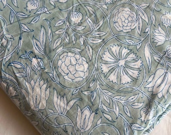 Op de afbeelding: Een gevouwen textiel met een licht saliegroene achtergrond en een herhalend patroon van witte bloemen en knoppen, omlijnd met dunne blauwe lijnen. Het ontwerp bevat ingewikkelde kronkelende stengels en bladeren die de bloemelementen verbinden.