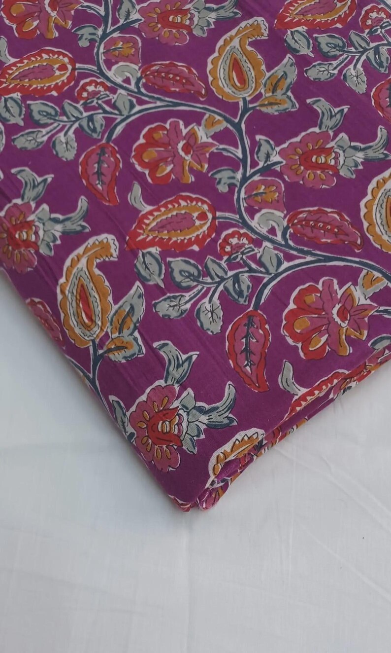 May include: A folded piece of fabric with a vibrant floral pattern. The design features paisley and flower motifs in shades of orange, red, and pink against a deep purple background. Gray-green leaves and stems complete the design.