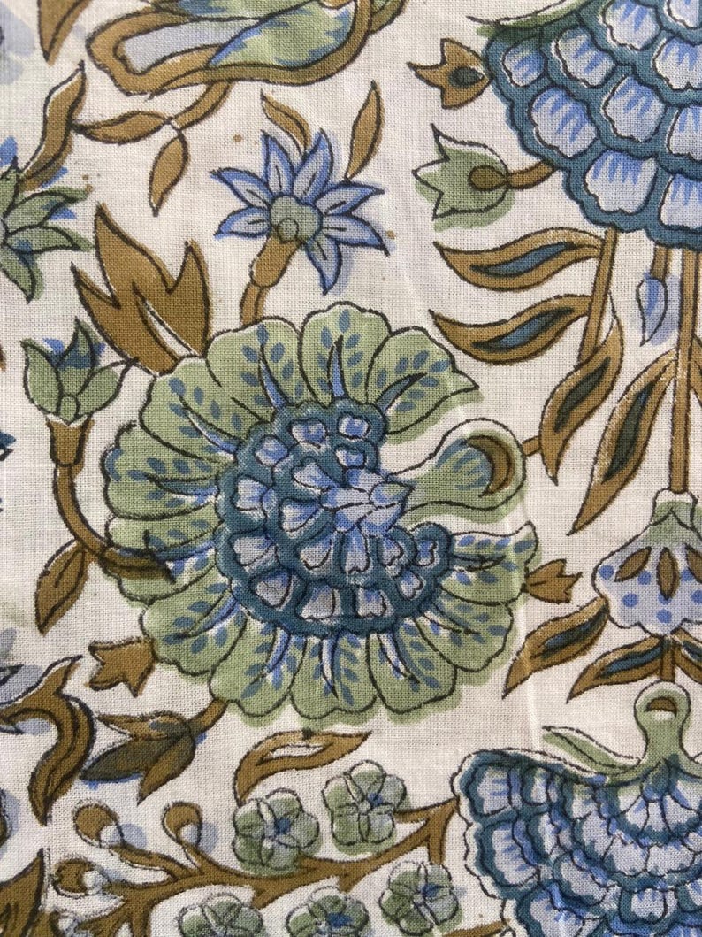 May include: Close-up of a fabric with a floral pattern. The design features large, stylized flowers in shades of blue and green, with brown stems and leaves, set against a cream-colored background. The fabric has a textured appearance.
