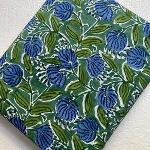 May include: A folded piece of fabric featuring a repeating botanical pattern. The design consists of blue, rounded, segmented flower blossoms and green leaves with darker green veins, all set against a solid medium green background.