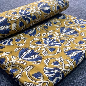 May include: Mustard-coloured fabric featuring a navy blue and white floral pattern. The fabric is folded and rolled, displaying the intricate design of flowers and leaves. The fabric is suitable for sewing or crafting projects.