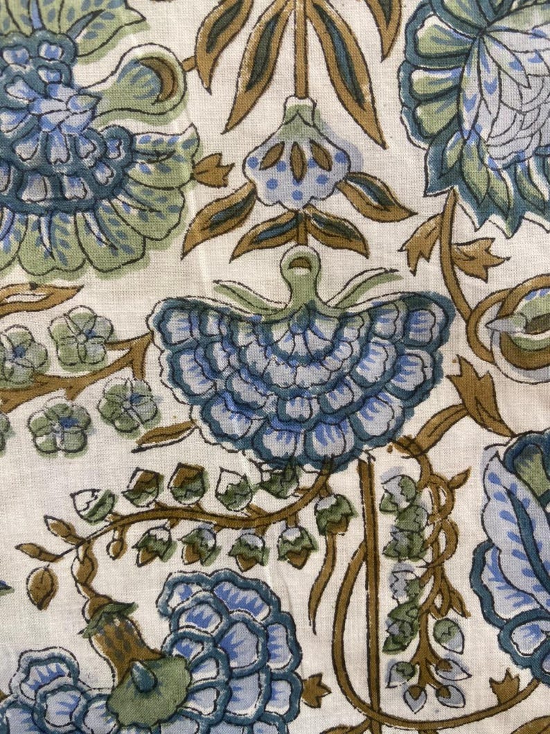 May include: Close-up of a fabric with a floral pattern. The design features large blue and green flowers with brown stems and leaves against a white background. The fabric has a vintage or bohemian aesthetic.