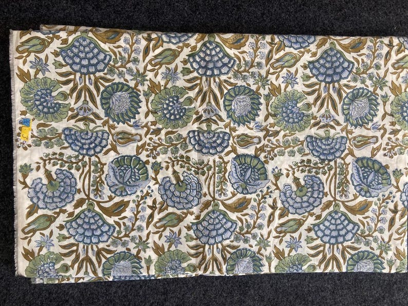 May include: A folded piece of fabric with a repeating floral pattern. The design features stylized blue and green flowers, brown stems, and small leaves on an off-white background. The fabric is likely cotton or linen.
