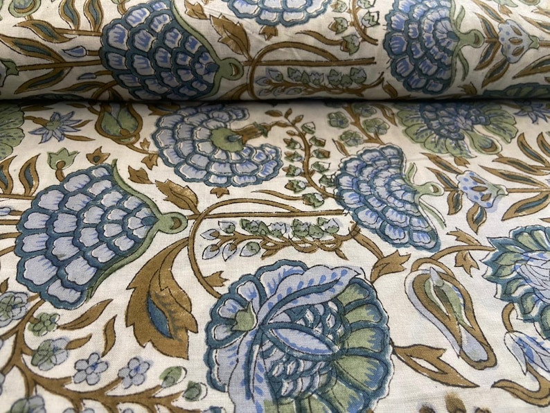 May include: Close-up of a fabric with a repeating floral pattern. The design features large, stylized blue flowers with green accents and brown stems and leaves, set against a cream background. The fabric is folded over.