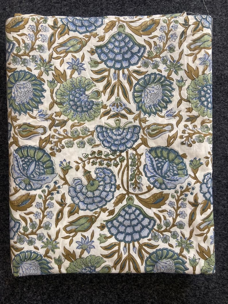 May include: A folded fabric with a repeating floral pattern. The design features large, stylized blue and green flowers with brown stems and leaves on a white background. The fabric is likely cotton or linen.